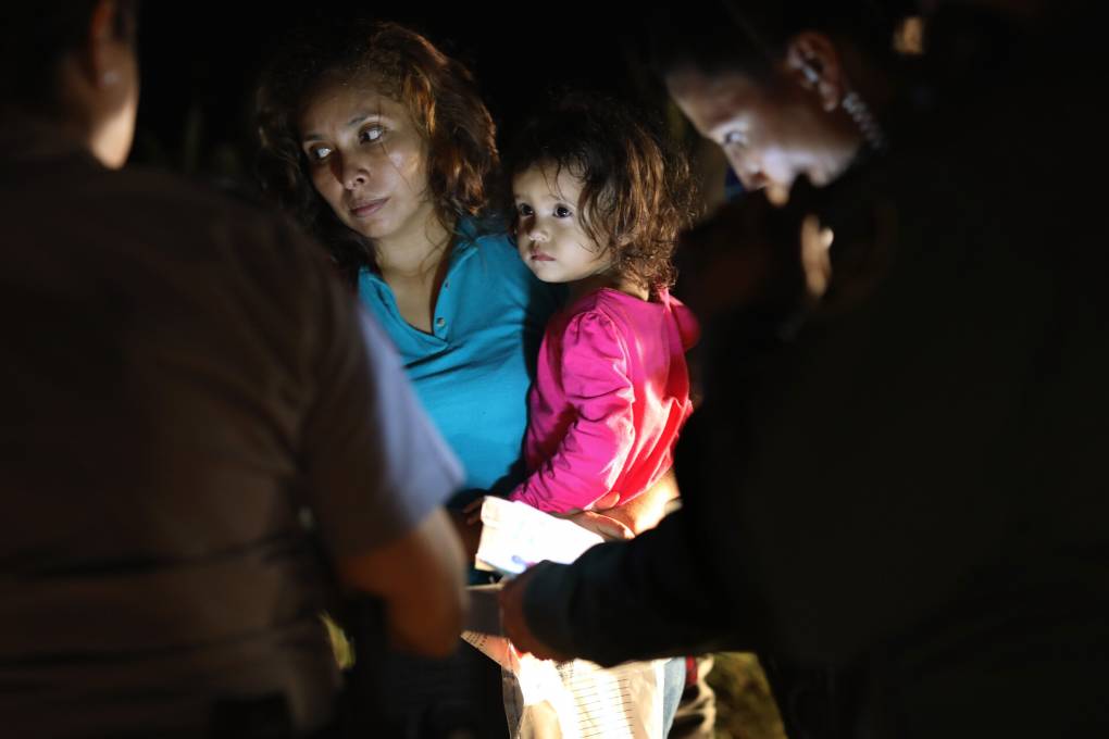 night-time shot of woman holding her small child, both with concerned faces, surrounded by silhouettes of border patrol agents
