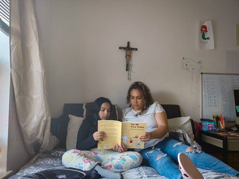 A mother and her young daughter read a book together on a bed, with a crucifix on the wall behind them.