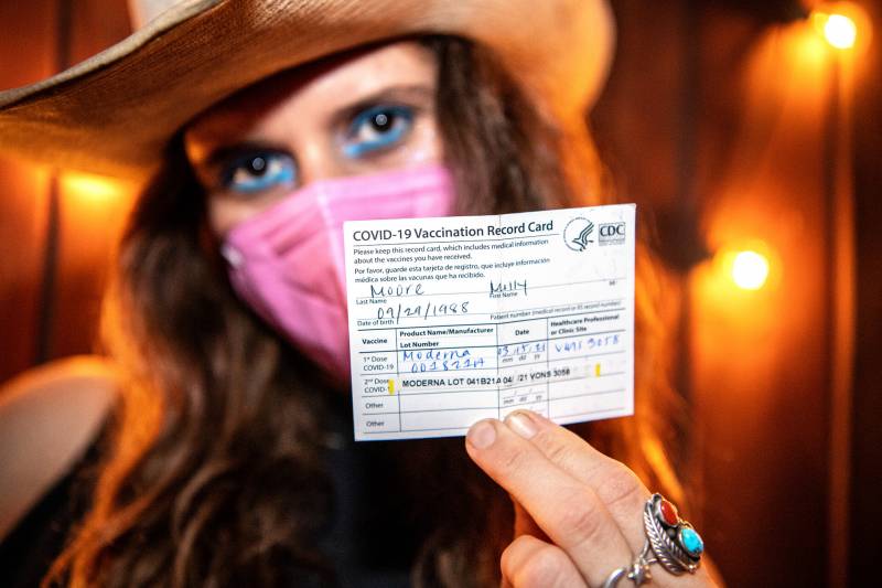 person wearing pink mask and cowboy hat with bright eyeliner holds up vaccination card