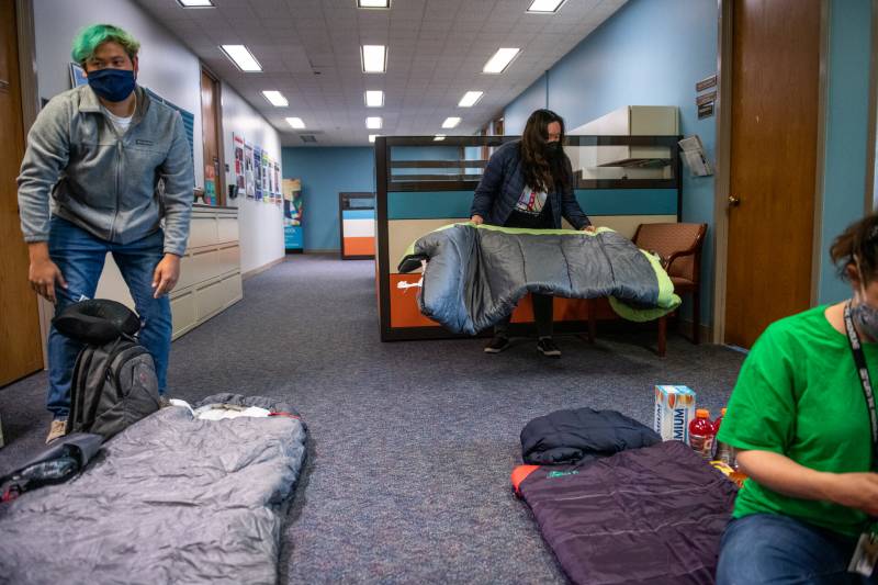 Three people lay down sleeping bags on the floor of an office.