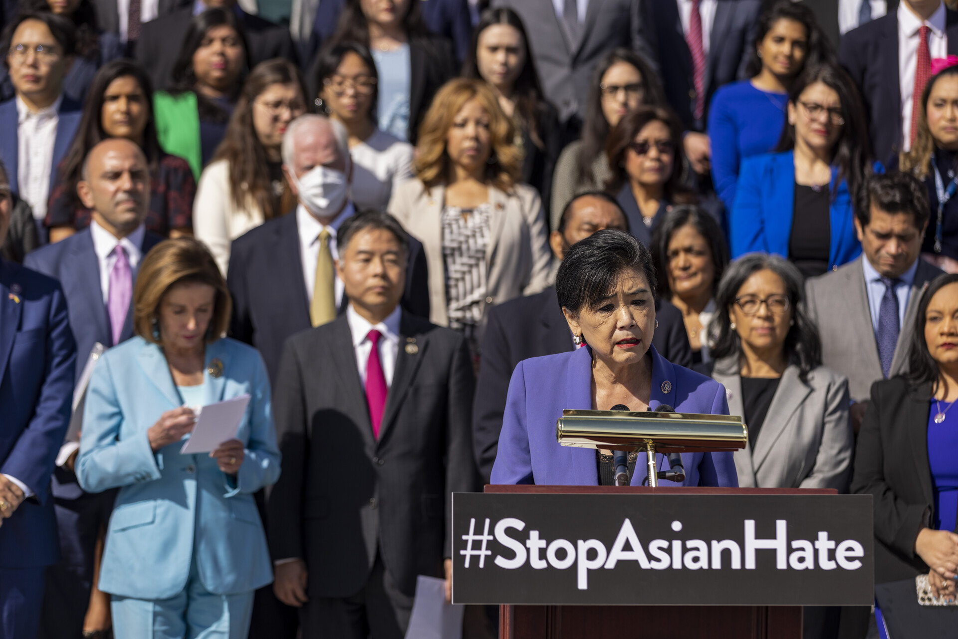 woman speaking at podium with 'stop asian hate' written on it as rows of faces of members of congress stand behind her