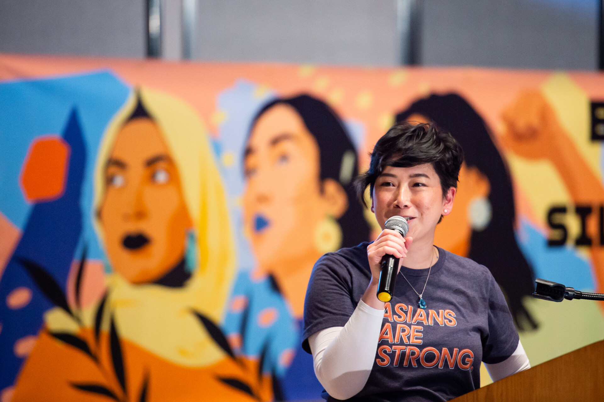 woman with 'asians are strong' t-shirt speaks at microphone with mural of strong female asian faces in background