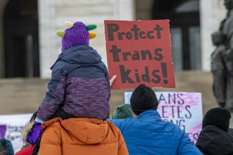 seen from behind, child in puffy coat sits on back of an adult, holding sign reading 'protect trans kids'