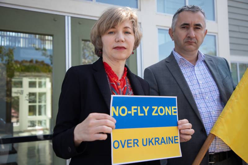 A woman and man stand outside an office building holding a Ukrainian flag and a sign that says, 'No-Fly Zone Over Ukraine.'
