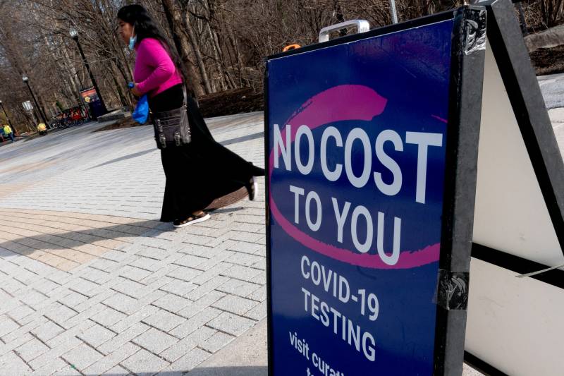 A woman wearing a pink shirt and a long flowing black skirt walks past a sign saying "no cost to you COVID-19 testing"