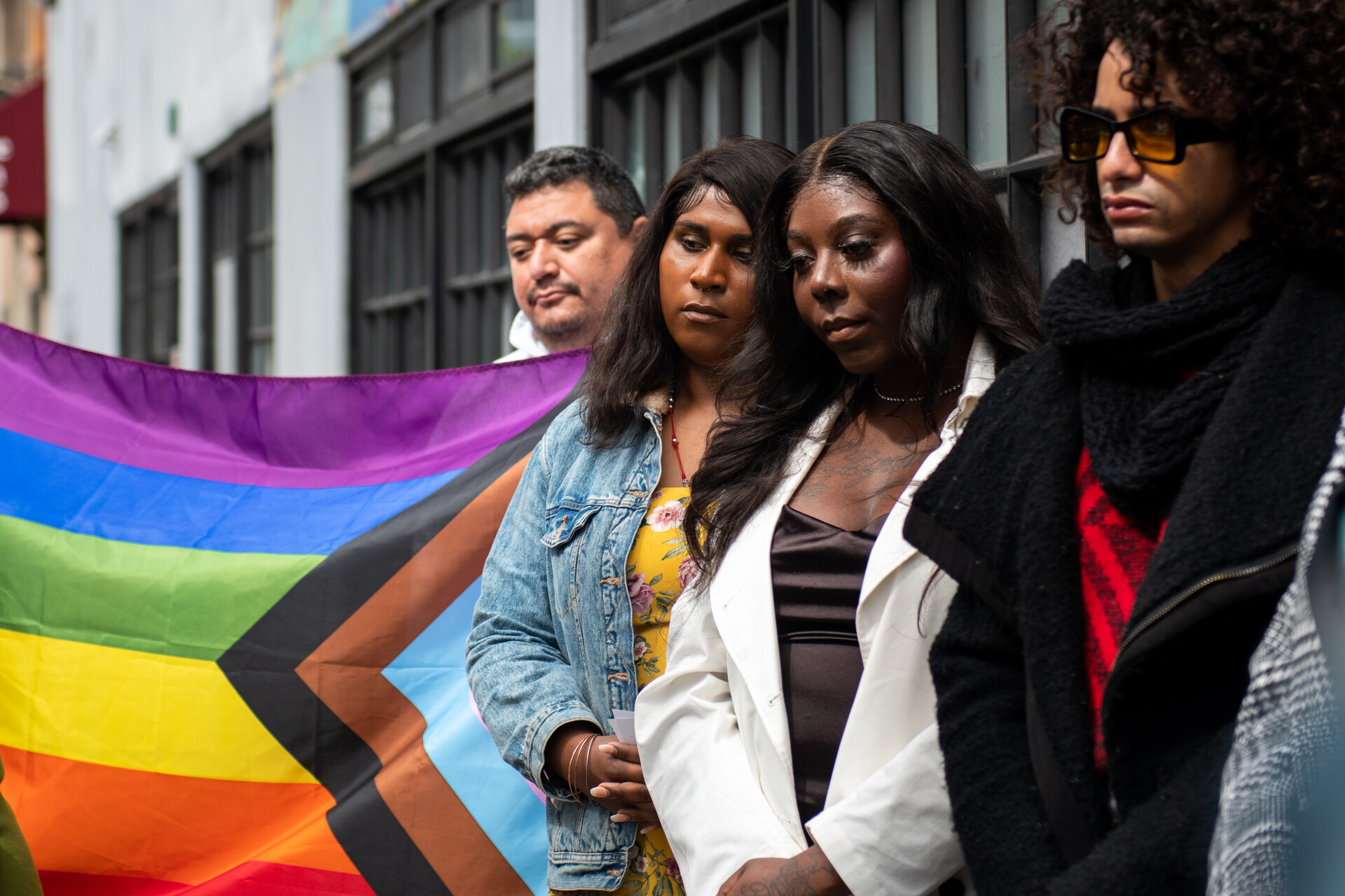 Black and brown people stand listening with stern faces as a pride flag is held in the background