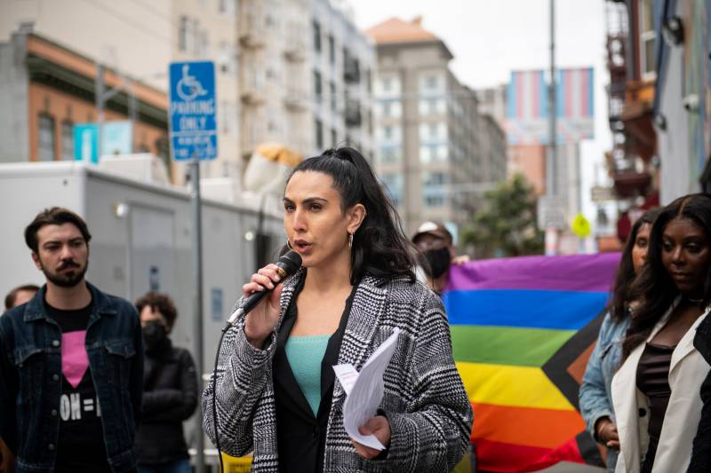 A person stands outside holding a microphone with other people standing around and holding a rainbow colored flag.