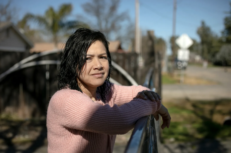 A woman wearing a pink shirt rests her arms on a fence outside.