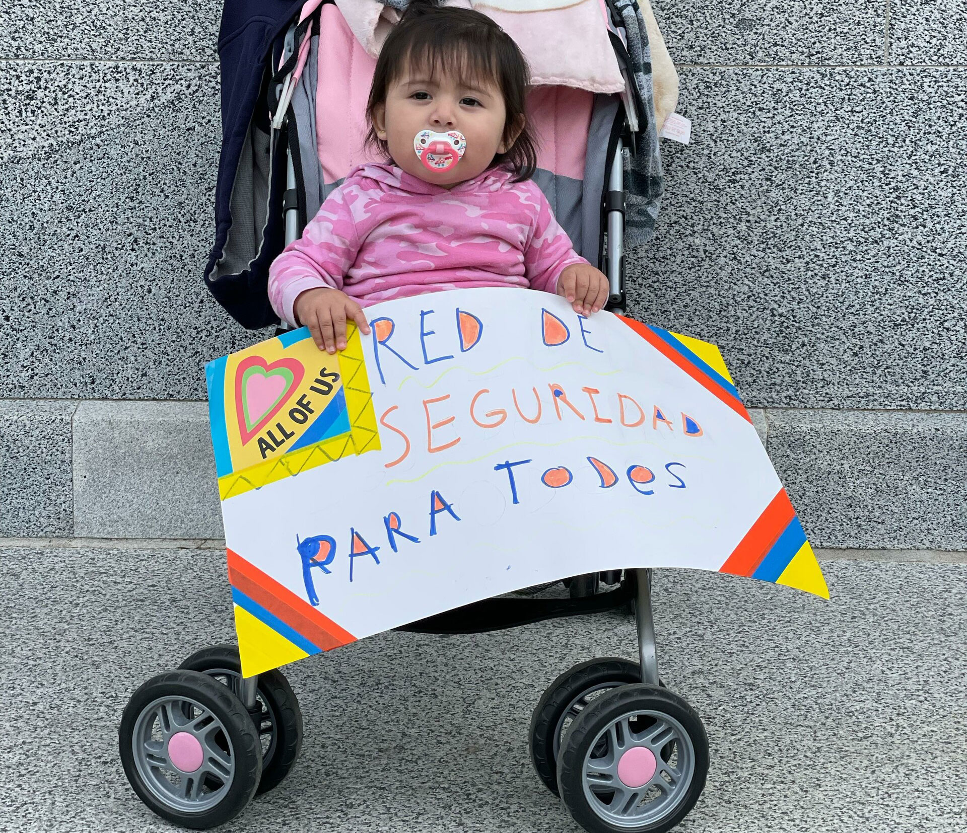 baby in stroller with pacifier in her mouth holds sign