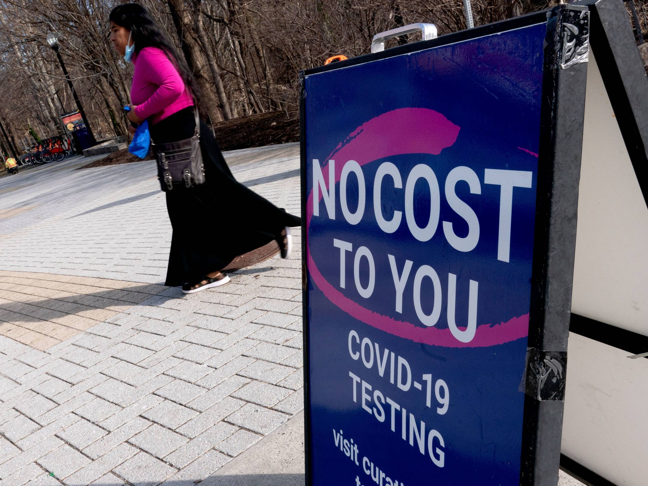 A woman with black hair and dark brown skin, wearing a black skirt and bright pink sweater walks across a stone plaza in the background. In the foreground is a blue sign saying "No Cost To You" COVID-19 Testing. A pink swirl wraps around the words: No Cost To You.