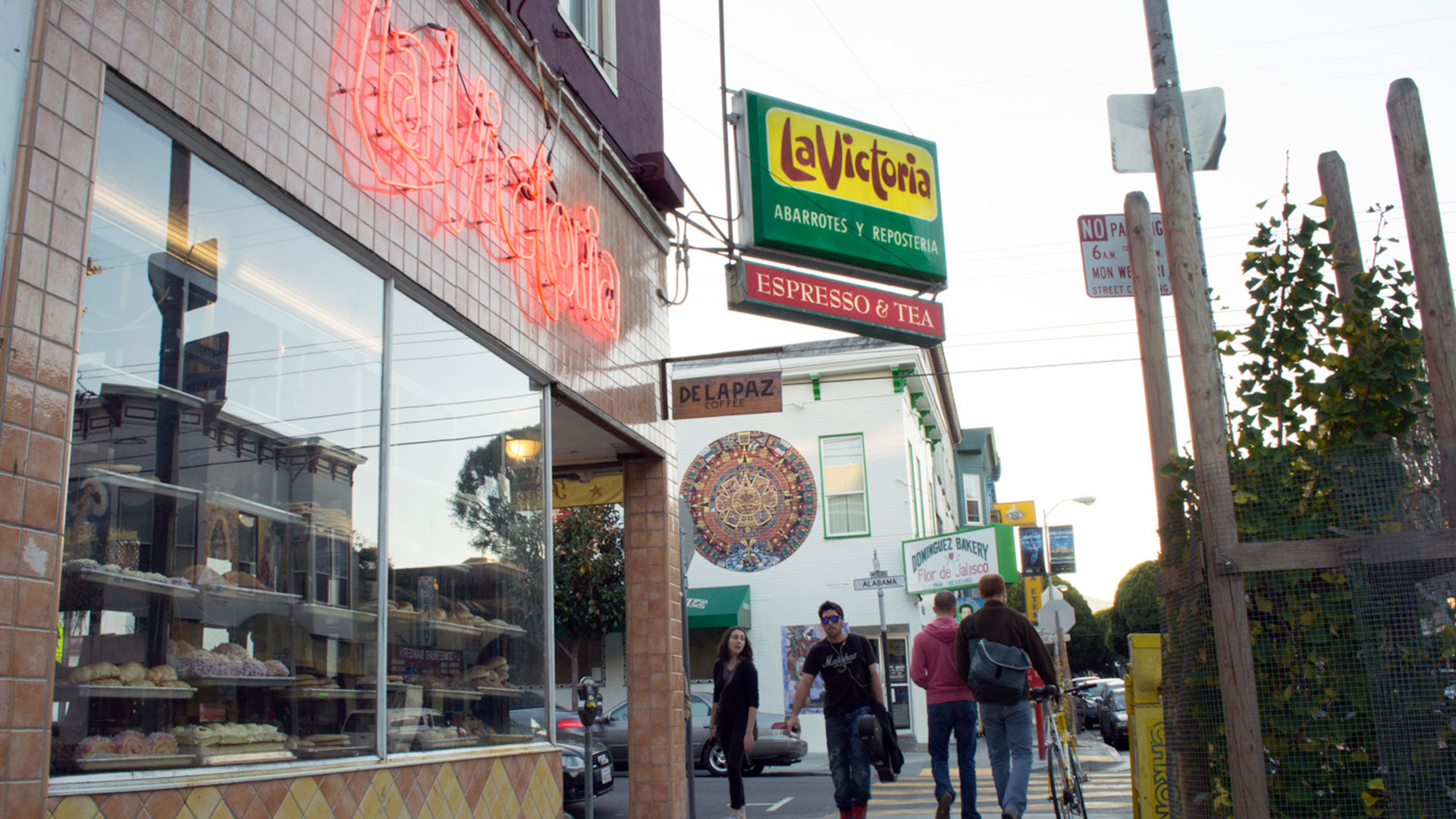 A neon sign reading La Victoria flickers above a glass window with rows of baked goods layed out behind it. A yellow, green and red sign sticks out from the corner of the building advertising abarrotes y reposteria.