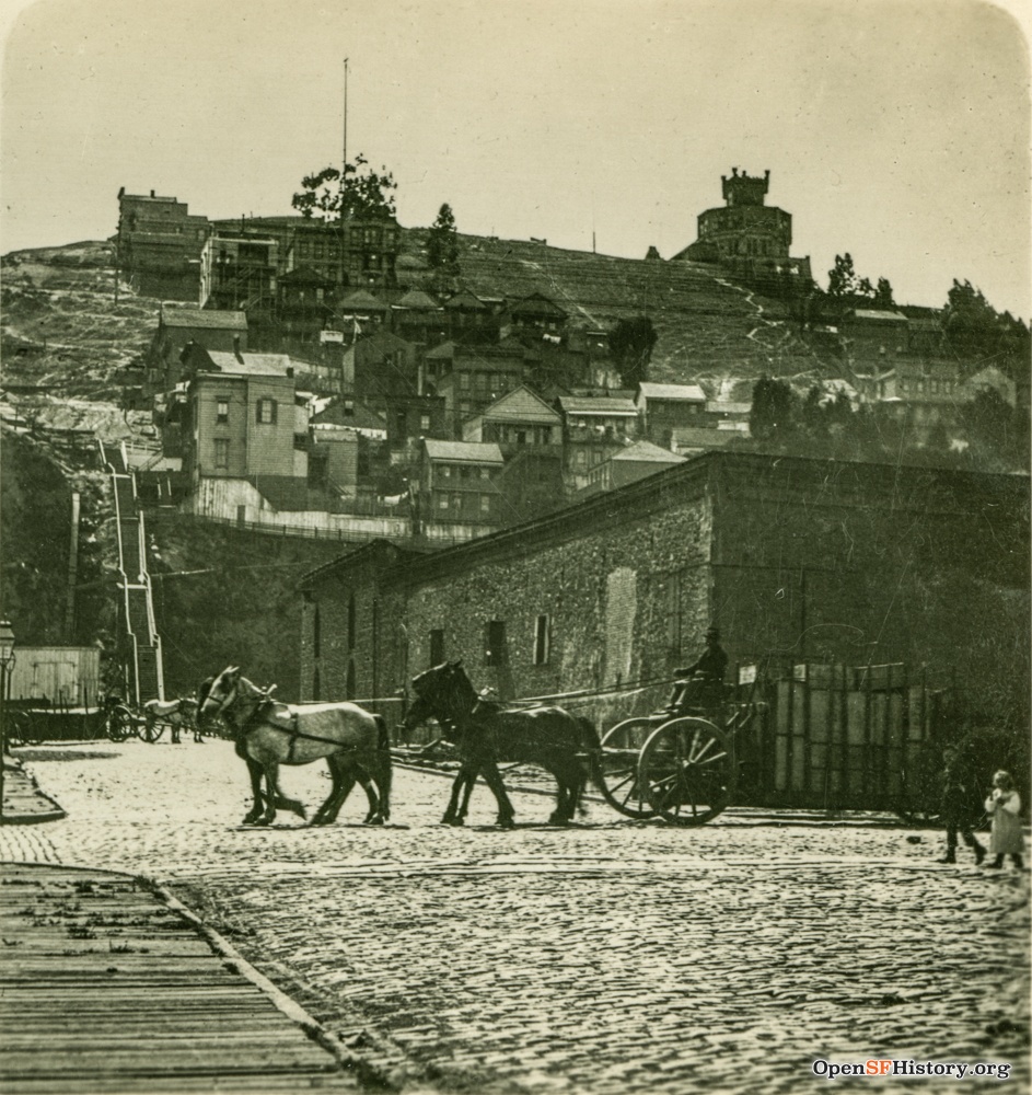 Black and white photo with horse and buggy in the foreground.  In the background, a wooden staircase climbs a steep, barren hill with a few low houses on it.