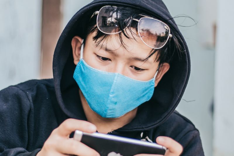 An Asian man wearing a black hoodie and a blue cloth face mask, with sunglasses balanced on the top of his head, gazes at a cellphone he holds in both hands
