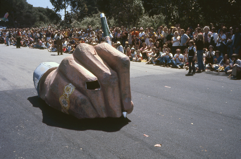What looks like a giant hand holding a pen rolls down a hill.  Spectators stand in the background