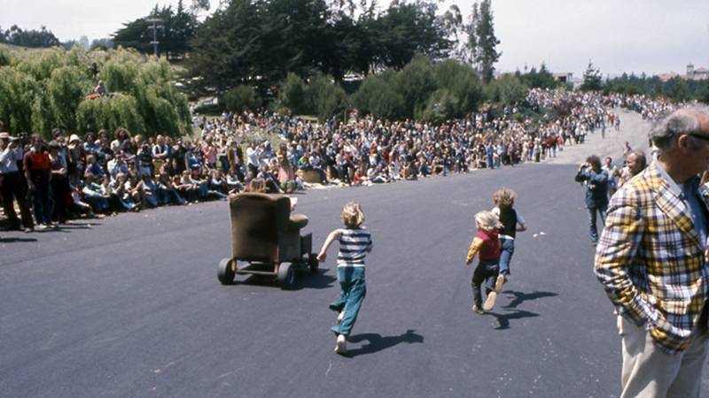 What looks like an armchair on wheels rockets down a wide steep street as kids in 1970s attire run after it. Along both sides of the road are throngs of spectators.