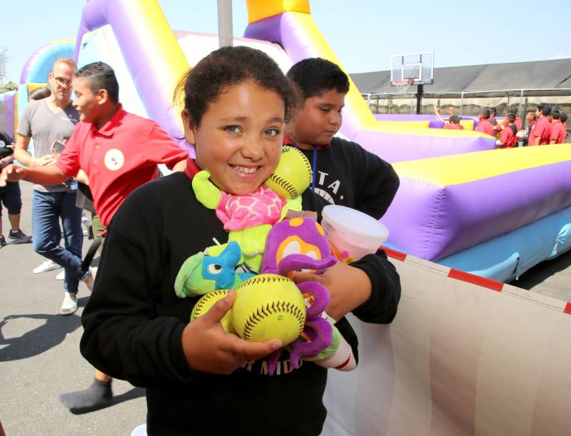 A middle-school-aged girl with brown skin and black hair smiles as she holds in her arms and tucked under her chin three yellow softballs and an assortment of small stuffed animals in yellow blue, green and purple. She's wearing a black sweater over a red shirt. She's standing next to a large blow-up slide in purple and yellow. This is a charity event for her school, Backpack Middle School.