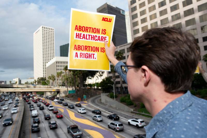 A white person with short, brown hair and black-framed glasses, dressed in a blue, patterned collared shirt, stands above a freeway holding out a yellow sheet of paper toward at least 10 lanes of traffic below. The paper says, “ACLU. Abortion is healthcare. Abortion is a right,” in bold, capitalized, red text. There are skyscrapers and palm trees alongside the freeway to the right, and at eye level, above the freeway, are at least two other pedestrian or car bridges.