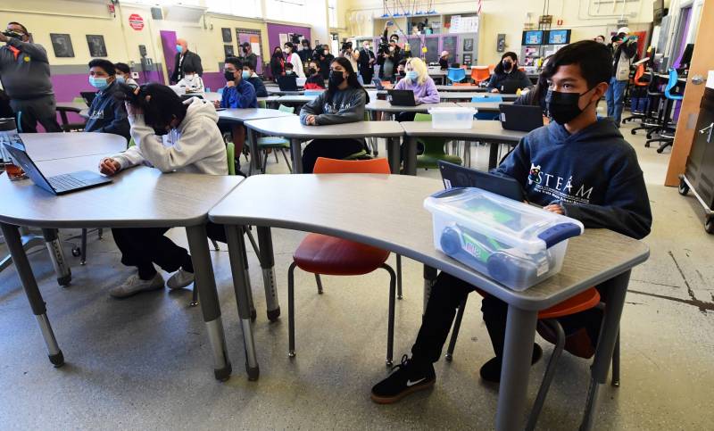Students wearing masks sit in a classroom with laptops open in front of them.