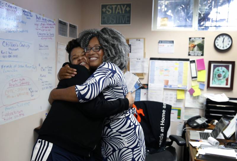 A teacher and student with wide smiles give each other a big hug in an office with a calendar, whiteboard and paper notes covering the walls. The teacher has black skin and is wearing a black and white dress in a graphic print. The student, almost as tall as her teacher, has black skin and is wearing a black sweatshirt and pants with white stripes down the side. 