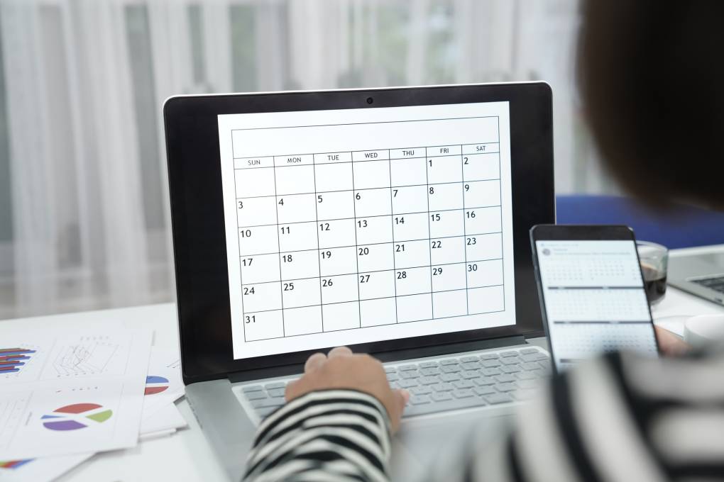 A person's hand is on a laptop keyboard that displays a calendar on a desk.