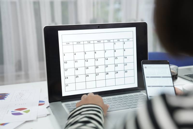 A person's hand is on a laptop keyboard that displays a calendar on a desk.