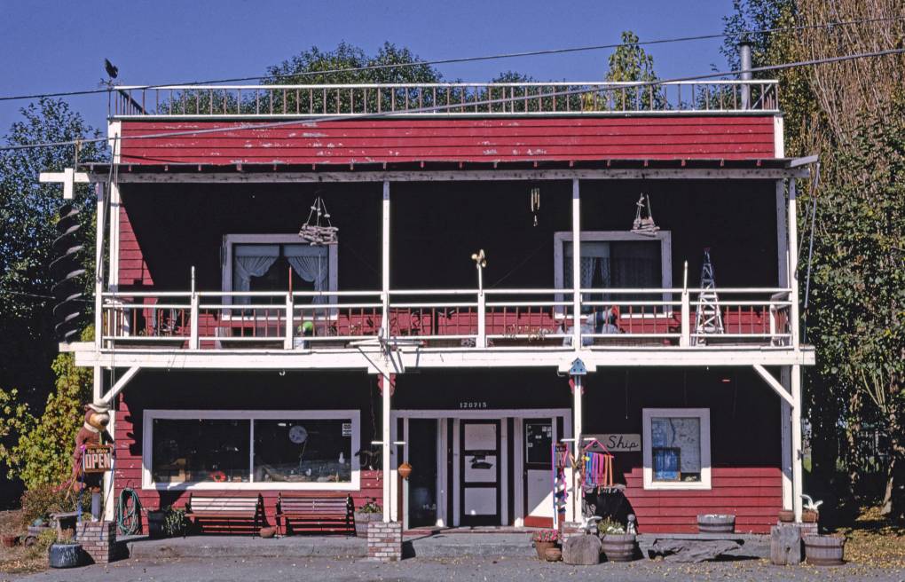 The general store in Orick, California is a two-storey building with wood siding painted in red, and battered with age. Four white poles run from the ground up through the second-story deck, with white trim along the deck. Lace curtains hang in the second-storey windows.