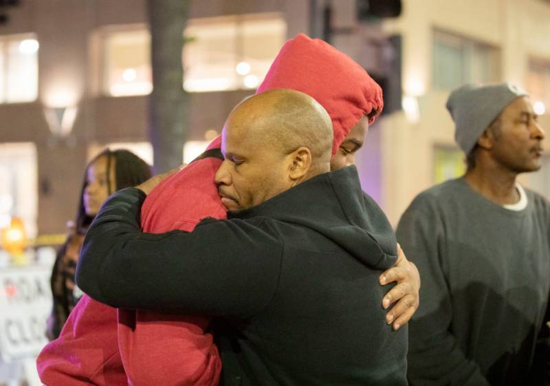 A man wearing a black hoodie embraces another person wearing a red hoodie pulled over his head with other people in the background.