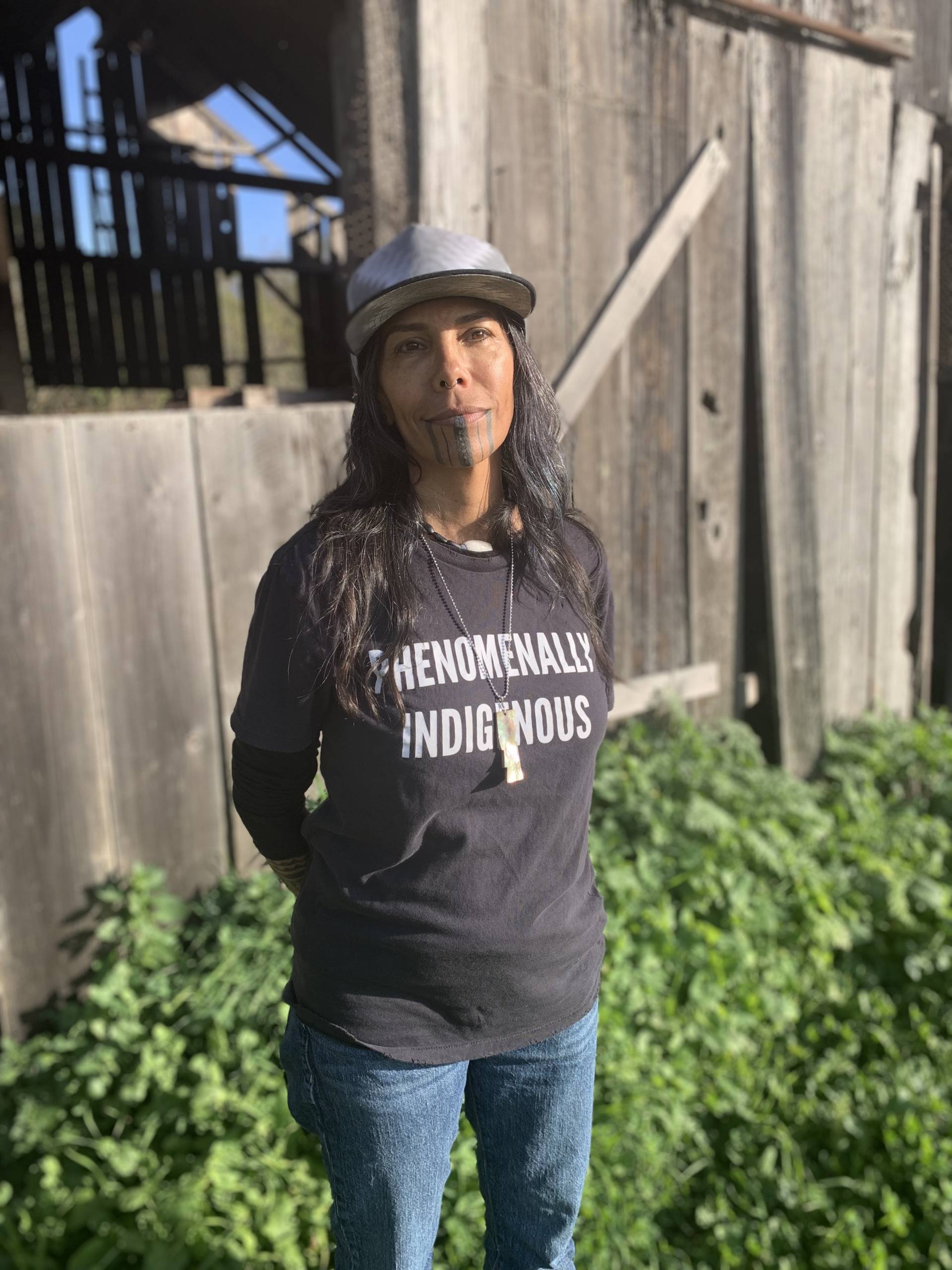 A women with a cap standing outside next to a barn, and wearing a t-shirt that says 'Phenomenally Indigenous.'