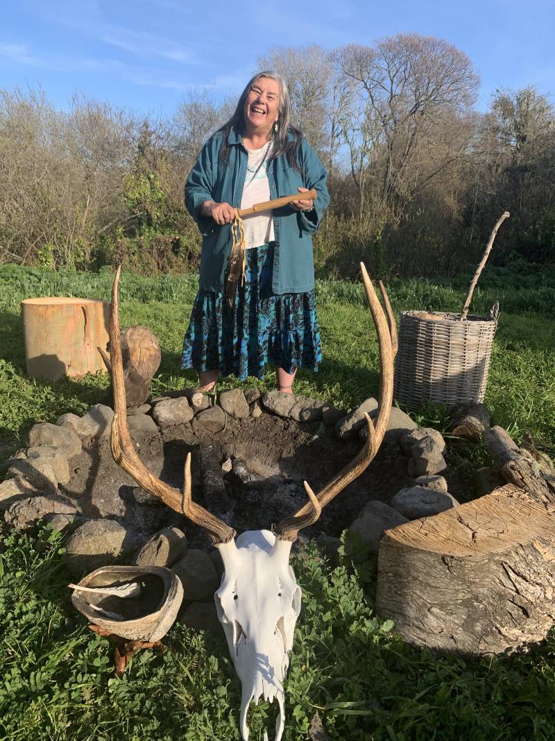 A woman holding a flat stick standing in field, behind the skull of an elk.