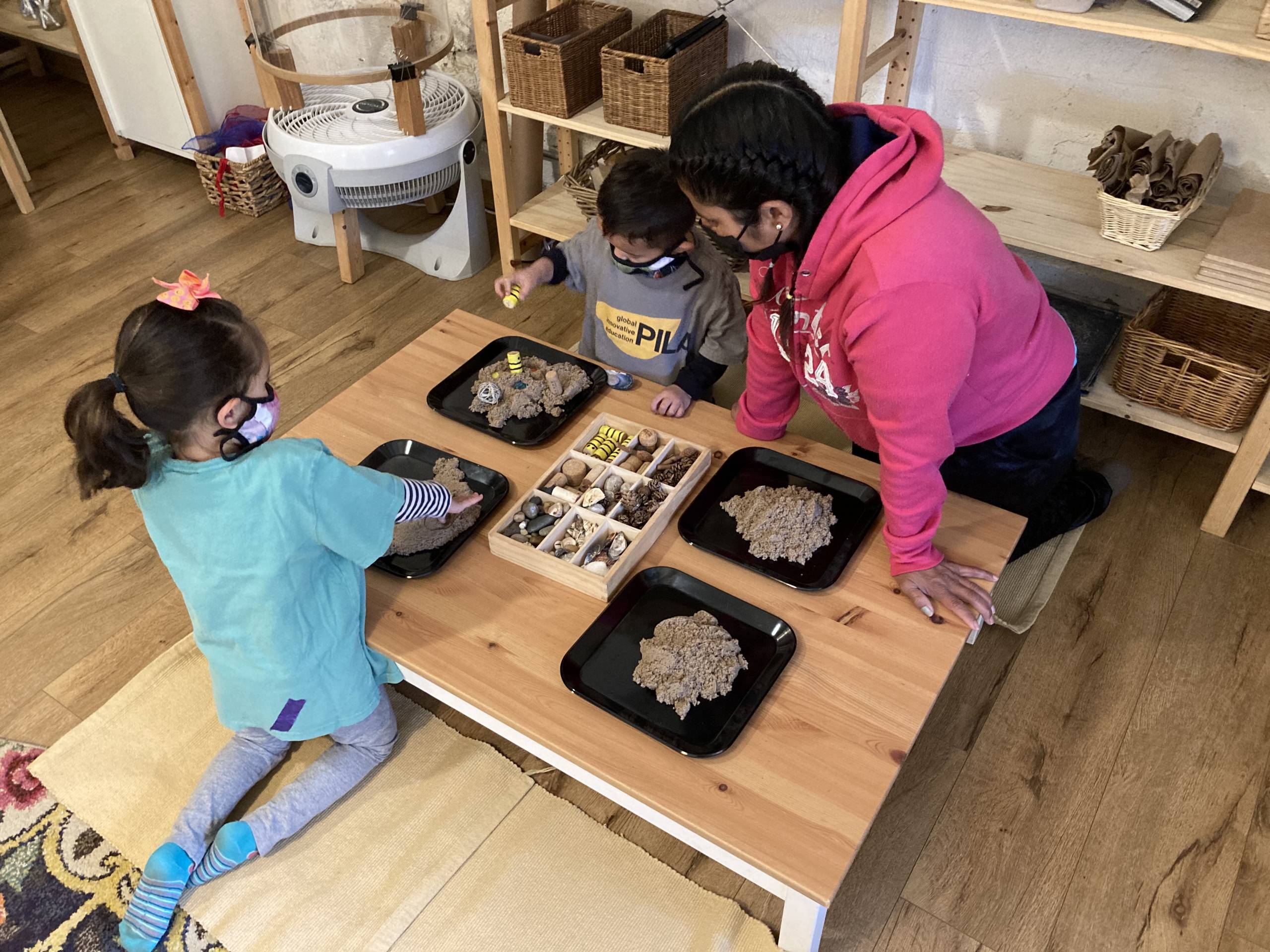 Two young students play with clay at a table, as a teacher supervises.