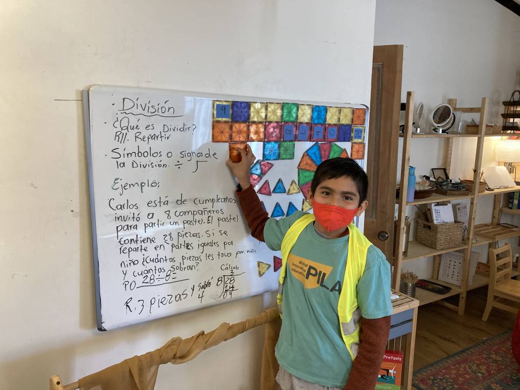 A young boy wearing a face mask points to a white board with writing in Spanish and shapes.