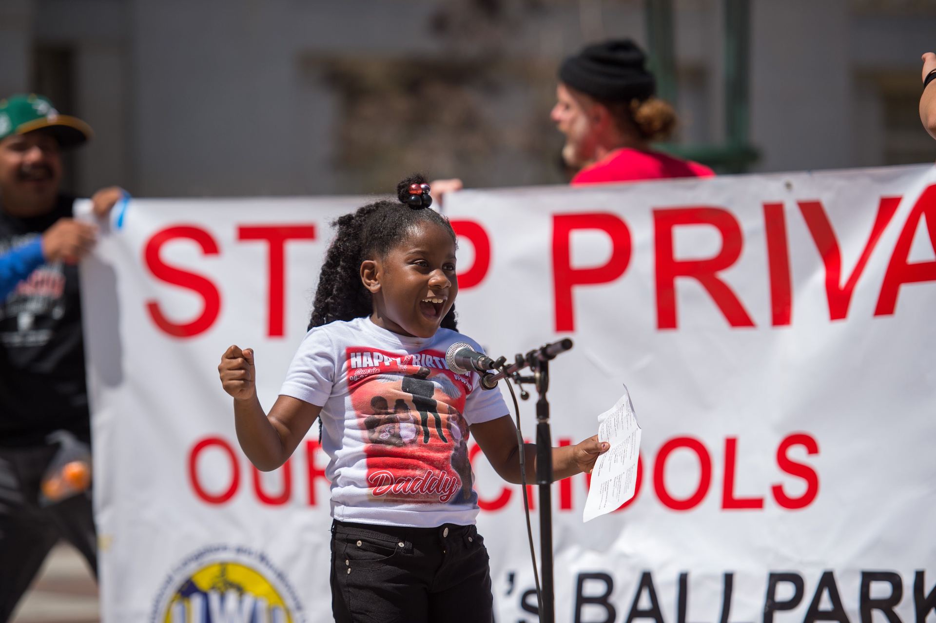 A young girl speaks into a microphone in front of a large banner.