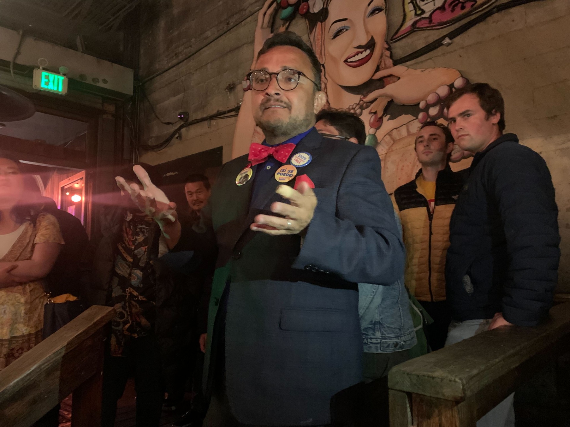 David Campos as seen from the waist up, hands outstretched, in a suit, speaking to supporters in front of a mural in a bar.