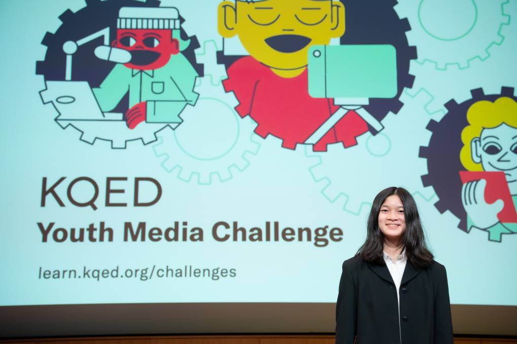 In the lower right of the frame stands a teenage Asian girl, smiling at the camera, with shoulder-length black hair and a black blazer over a white blouse. Most of the photo is taken up by the digital screen behind her, which shows three cartoon graphics of a person either speaking into a radio, standing behind a camera, or reading a script, inside a border/outline that looks like a gear. In the lower left are the word "KQED/Youth Media Challenge."