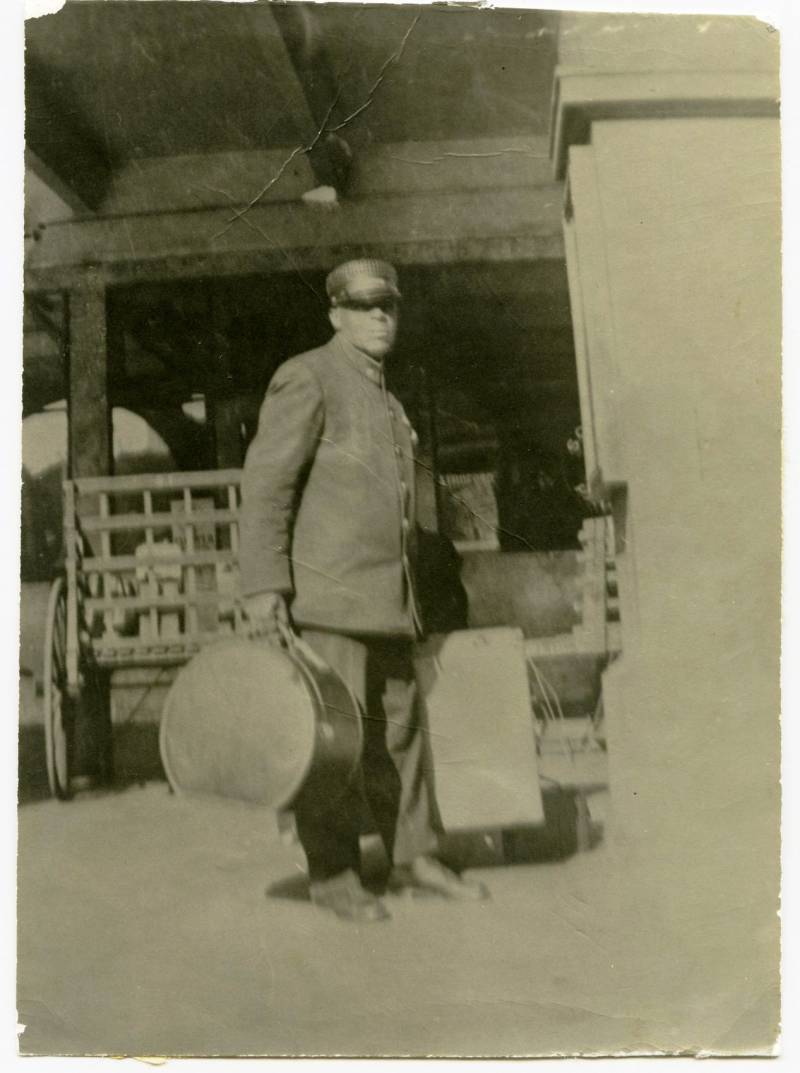 A very old and poor quality image shows a man wearing a pullman porters uniform holding 2 pieces of luggage at a train station.