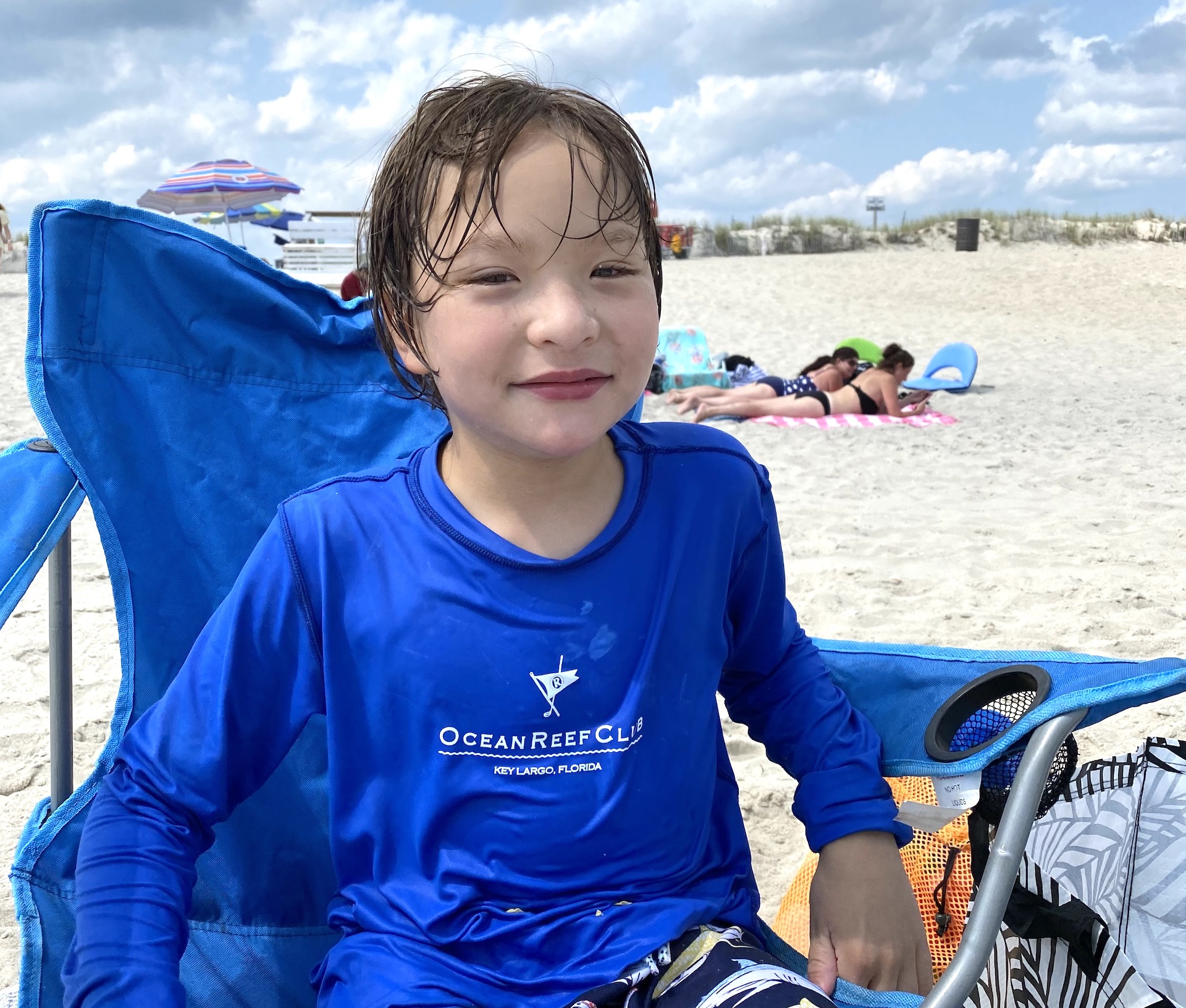 A young boy wearing a swim shirt and swim suit sits in folding chair on a beach.