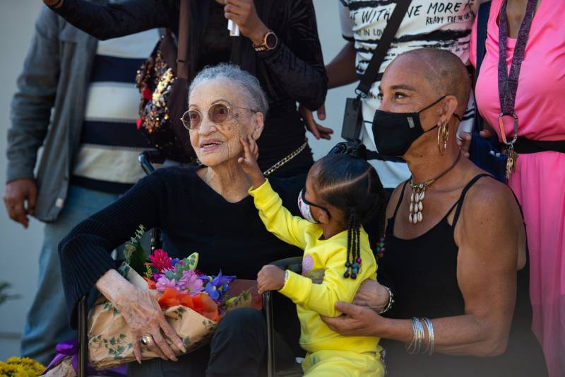 An elderly woman sits holding a bouquet of flowers, as a little girl touches her face.