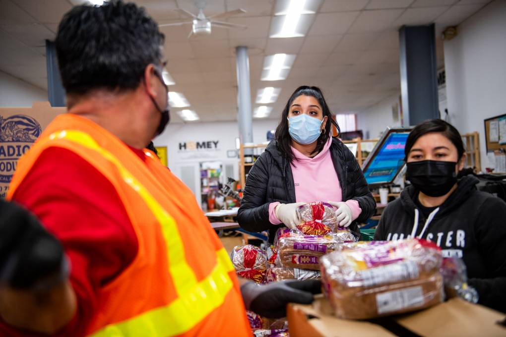 Gabriela Alemán, co-founder of Mission Meals, works alongside a group of volunteers to pack bread, fruit, vegetables, eggs and milk into boxes. A young Latina woman, she is wearing a pink shirt with a black puffy jacket over it, and looking over at a man wearing a neon orange work shirt in the foreground of the image.