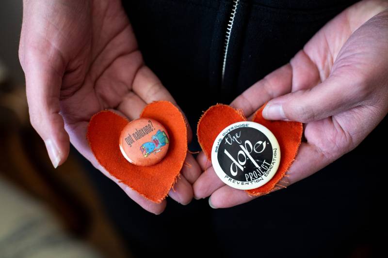 Two hands hold orange hearts with pins on them. One pin is orange and says, "got naloxone?" The other is black with a white border and reads, "The Dope Project."