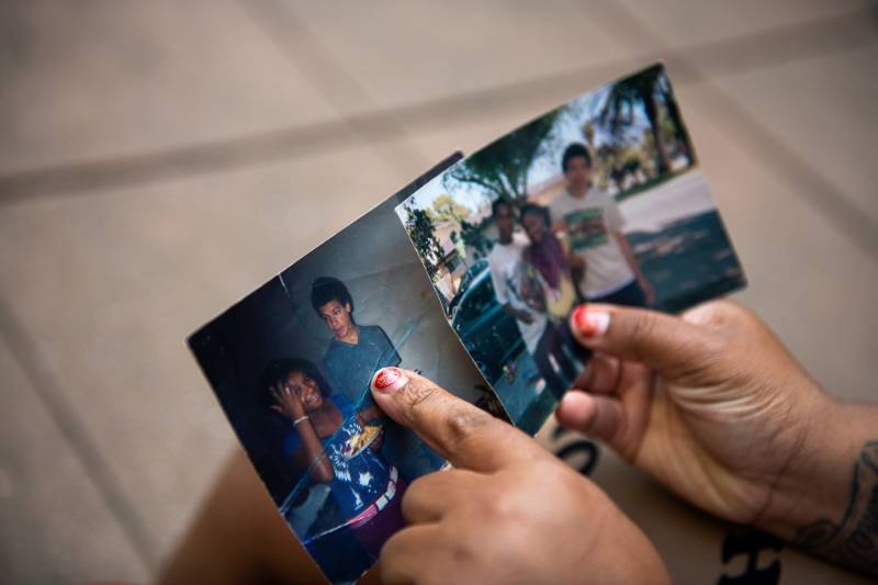 Hands with chocolate skin and chipped red fingernail polish hold two photos. One photo shows the woman and her cousin as teenagers. In the photo, her cousin appears male, with dark hair and a blue shirt. The other photo is a blurry image of three young people in dark pants and white shirts.