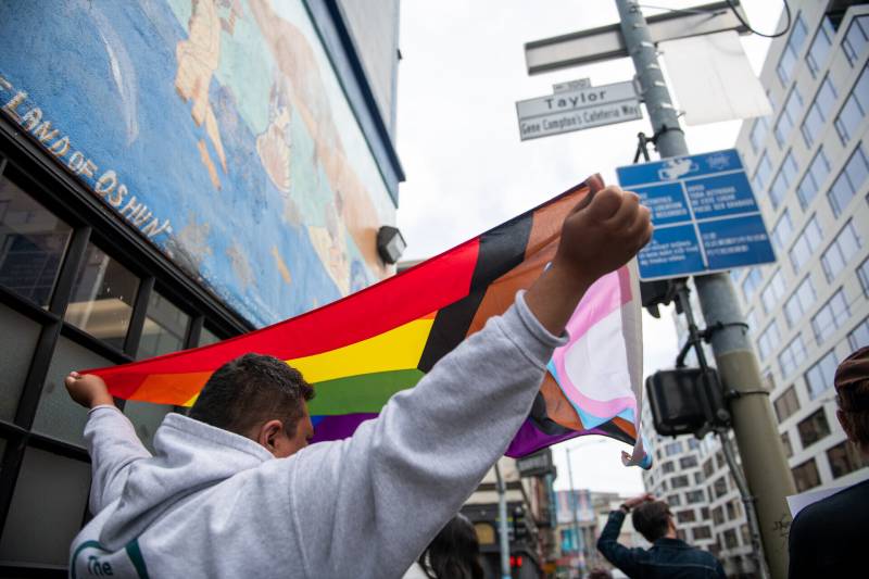 A man on the street hold a multi-colored flag above his head.