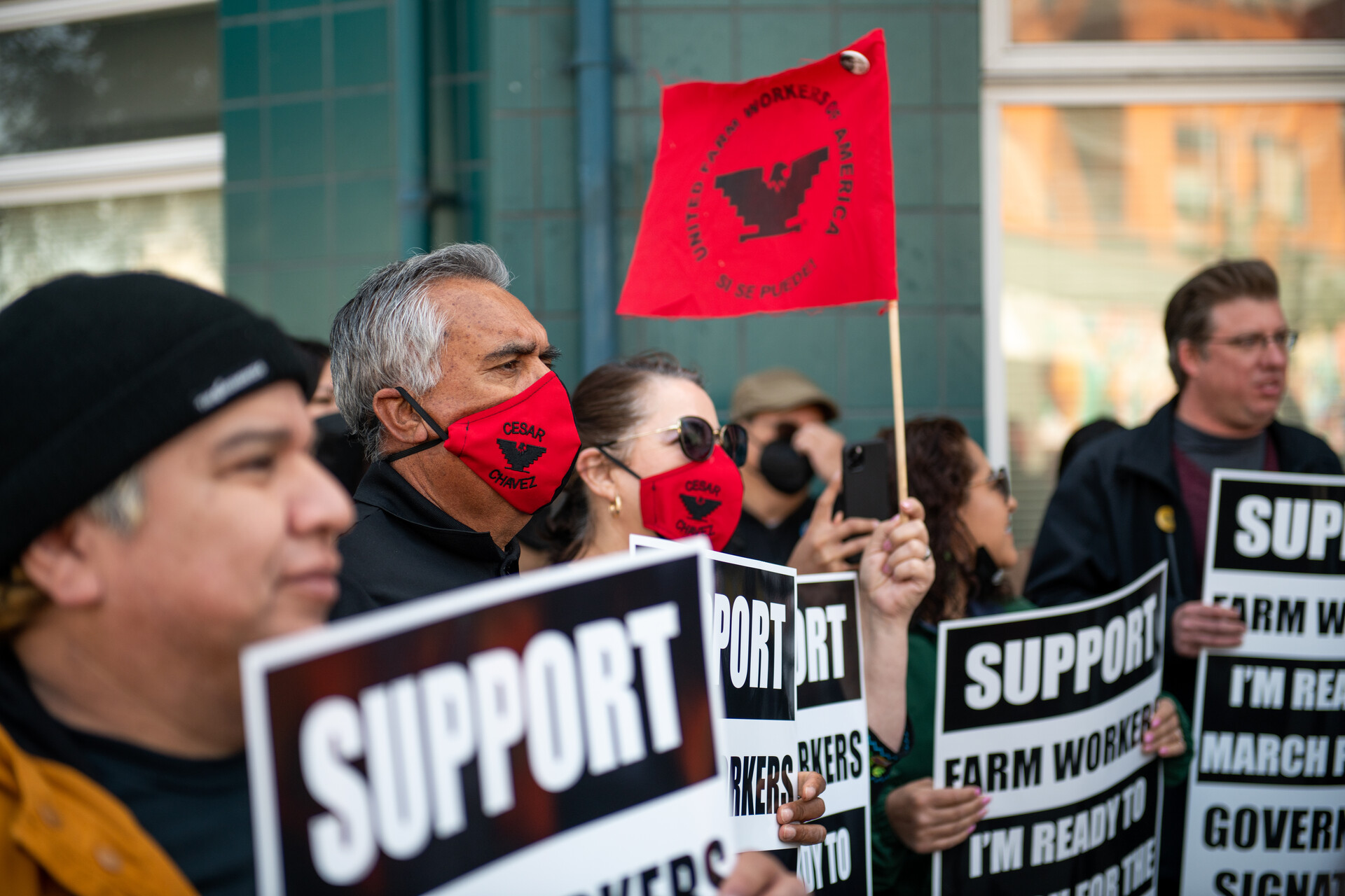 A man and a woman in focus amid other demonstrators, slightly blurred, wear red facemasks reading 'Cesar Chavez' in black with the black UFW phoenix symbol, and hold signs black-and-white signs reading 'Support Farm Workers: I'm ready to march for the governor's signature.'