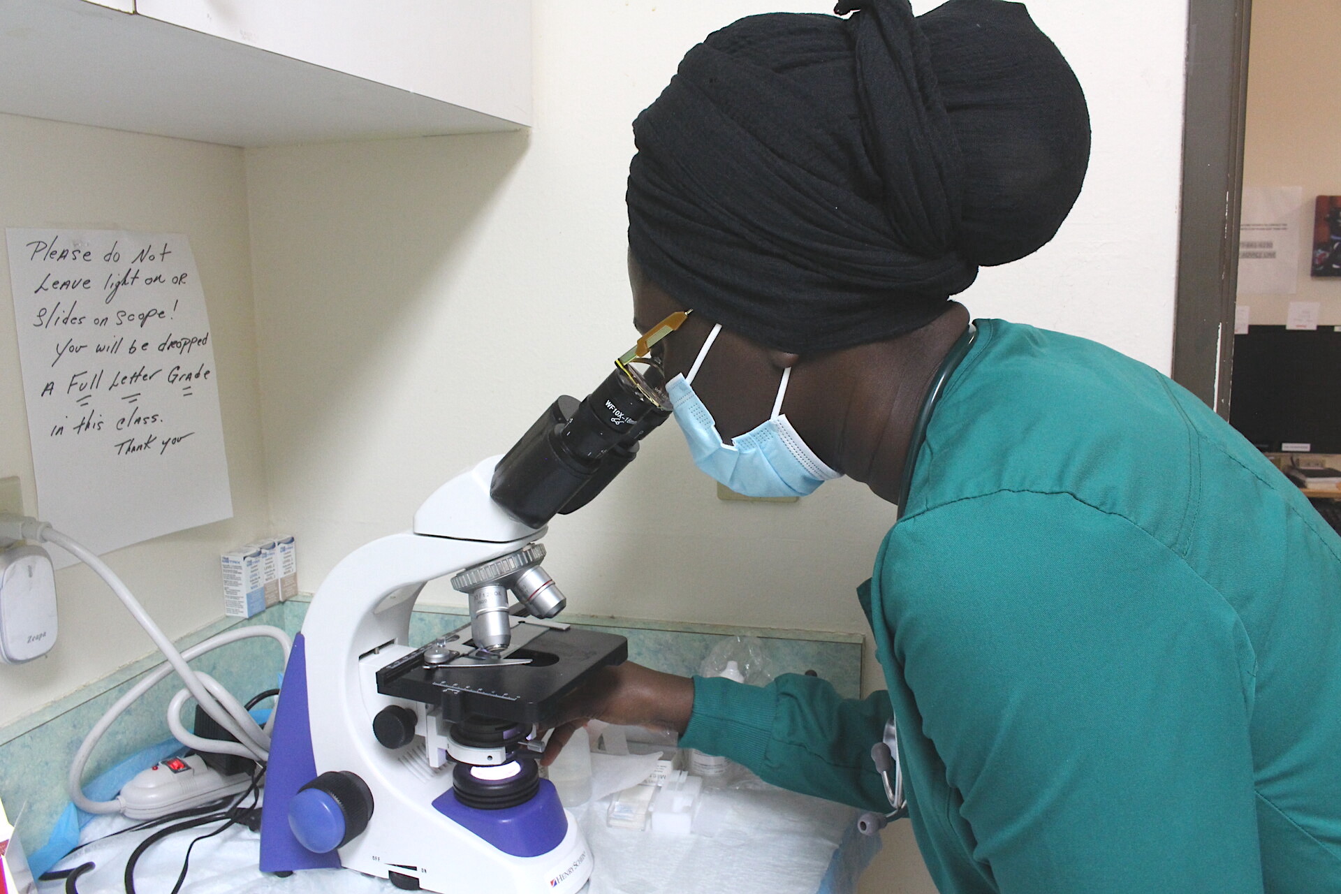 A woman in green medical scrubs looks into a microscope.