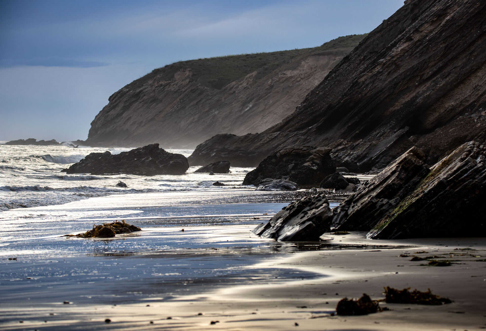 A view up a sandy shoreline alongside steep, shaded, rocky cliffs, with the sun shining on receding waves.