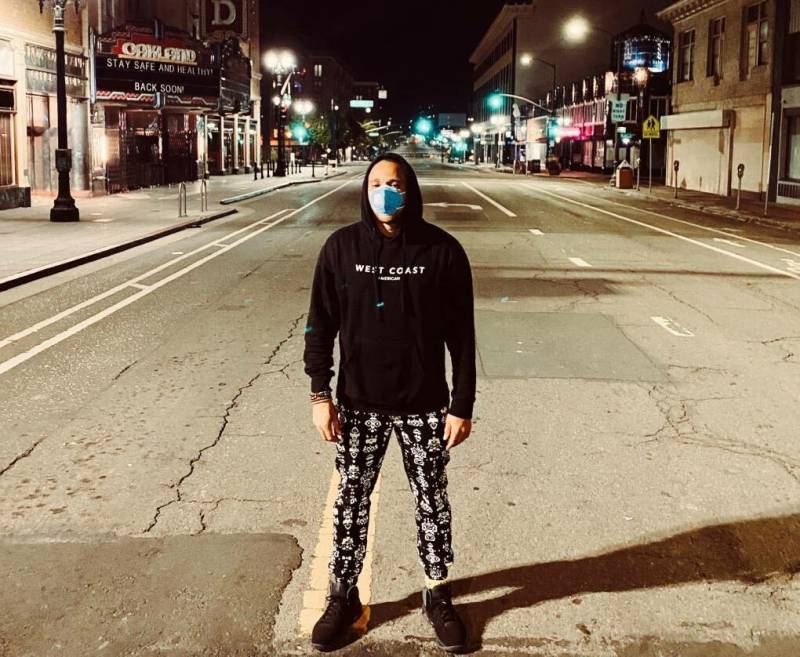 A man wearing a black sweatshirt with the hood pulled up over his head, a powder-blue face mask, black-and-white-patterned pants, and black shoes, stands in the middle of a cracked city street lit by what seems like a streetlight just to his left. Additional streetlights and neon signs illuminate the street and shops behind him. No cars are parked anywhere or appear anywhere on the street.