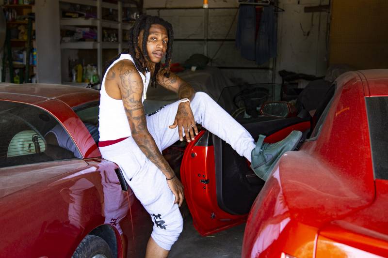 A Black man with shoulder-length locks wearing a white undershirt and white pants hitched to below his knees, with tattoos on both arms, both hands, his back and the contours of his face, leans against one red sports car inside a garage, with his left leg (and a black-sneakered foot) propped on a second red sports car, whose door is open. He looks directly at the camera. Beyond the cars in the half-dark, shelves are visible, some empty and some holding what might be cleaning supplies or extra food supplies.