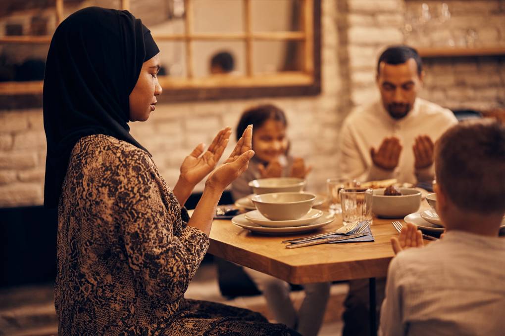 A Muslim family sits around a dining table, eyes closed, palms lifted face-up. On the left, at the head of the table, sits a woman wearing a black hijab and a patterned dress. Two children sit on either side of her with their hands in the same position; we see the back of a small, close-cropped head to her right, and to her left sits a young girl with shoulder-length dark hair, smiling with her eyes closed. Beside the girl sits a man wearing a white button-up shirt. The table is set neatly with white bowls and plates and clear glasses. The wall behind the table is white-painted brick, inset with a wall-size mirror. The overall tones in the photos are cream and sepia.