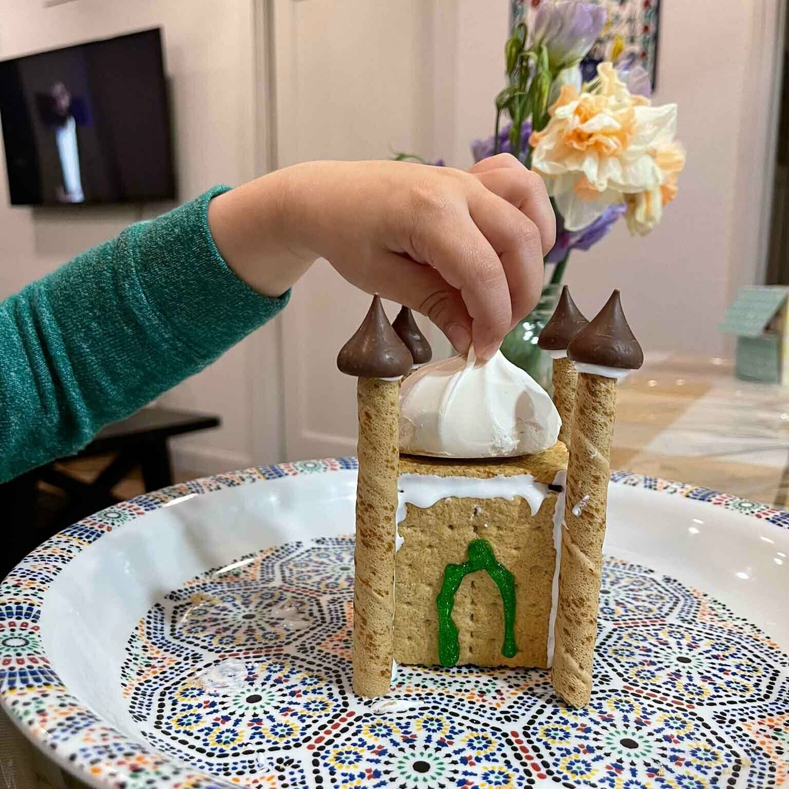 A child's hand gently holds an edible mosque structure about 5 inches high, constructed of graham crackers and minarets made of chocolate, standing on a table 