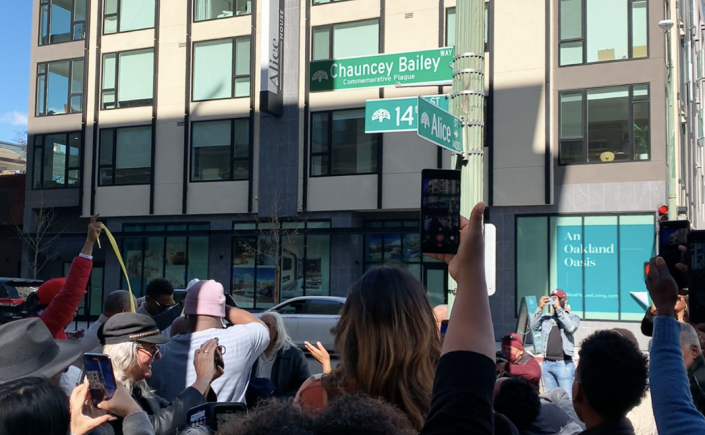 A crowd of people face a street sign with their cell phones in the air.