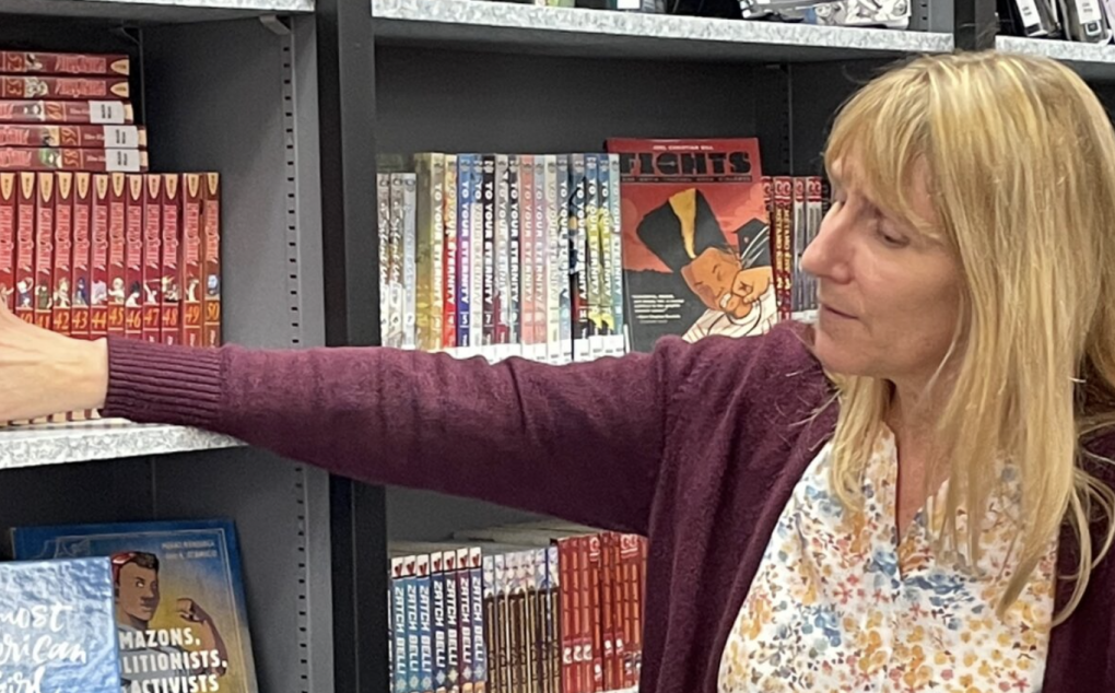 A woman wearing a reddish sweater and light shirt points towards books on a shelf.